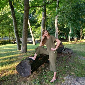 May include: A woman wearing a green linen jumpsuit with short sleeves and wide legs, sitting on a fallen log in a forest setting.