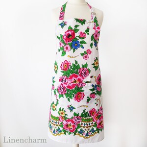 May include: A white apron with a floral print featuring pink, red, blue, and yellow flowers. The apron has a large pocket on the front and a tie at the neck.