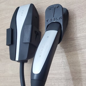 May include: Two black Tesla charging adapters with a silver connector and a black cable. The adapter on the left has a white label with the word "Tesla" printed on it. The adapter on the right has a black label with the word "Tesla" printed on it.