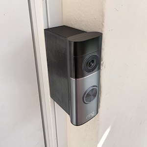 May include: A black and silver Ring video doorbell mounted on a white wall. The doorbell has a camera lens, a button, and the Ring logo. The mounting bracket is black and rectangular.