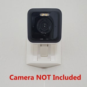 May include: White wall mount for a security camera. The mount has a black camera housing with a lens and two small lights. The text "Camera NOT Included" is printed below the mount.