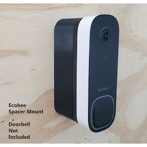 May include: A black and white Ecobee doorbell camera with a white trim and a black button. The camera is mounted on a wooden surface. The text "Ecobee Spacer Mount" is written below the camera. The text "Doorbell Not Included" is written below the text "Ecobee Spacer Mount".