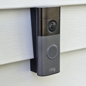 May include: A gray and black Ring video doorbell mounted on a white siding. The doorbell has a camera lens, a large circular button, and the brand name "ring" printed on the bottom.