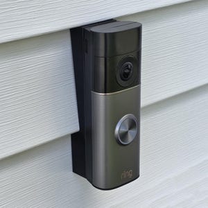 May include: A rectangular video doorbell device with a black upper section containing a camera lens and a metallic grey lower section featuring a circular button and the word Ring printed in white at the bottom, mounted on a white siding wall.