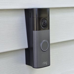 May include: A rectangular video doorbell with a black upper section containing a camera lens and a grey lower section featuring a circular button and the word ring printed in white. The device is mounted vertically on a white horizontal siding wall.