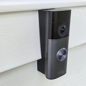 May include: A black and gray Ring video doorbell mounted on a white siding. The doorbell has a camera lens, a silver button, and the Ring logo. The doorbell is attached to a black mounting bracket.