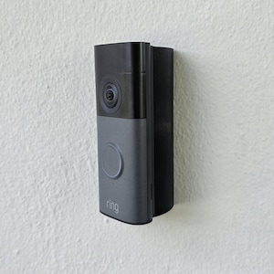 May include: A gray and black Ring video doorbell mounted on a white textured wall. The doorbell has a camera lens, a large round button, and the Ring logo. The device is rectangular with rounded edges.
