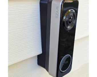 May include: A black and silver Wyze video doorbell mounted on white siding. The doorbell has a camera lens, a silver accent, and the Wyze logo. The doorbell is rectangular and has a sleek, modern design.