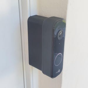 May include: A black smart doorbell with a camera and a large button, mounted on a white wall. The doorbell has the "myQ" logo on the bottom. The device is rectangular with rounded edges.