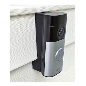 May include: A black and silver video doorbell mounted on a white siding. The doorbell has a camera lens, a silver button, and the brand name "ring" printed on the bottom. The doorbell is attached to a black mounting bracket.