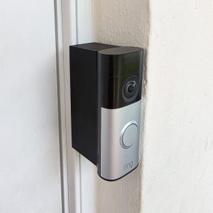 May include: A black and silver Ring video doorbell mounted on a white door frame and wall. The doorbell has a camera lens, a large silver button, and the Ring logo. The device is rectangular with rounded edges.