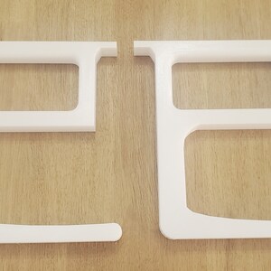 May include: Two white plastic shelf brackets, each with a rectangular opening and a curved bottom edge.