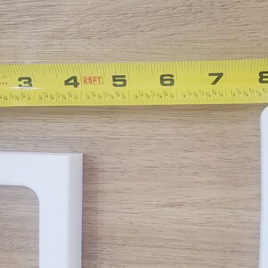 May include: A yellow measuring tape with black markings is placed on a light brown wooden surface. The tape reads "25FT" and shows measurements in inches. Two white plastic pieces are positioned below the tape.