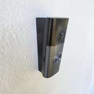 May include: A dark gray Ring video doorbell mounted on a white textured wall. The doorbell has a camera lens, a silver button, and the Ring logo. The device is rectangular with rounded edges.