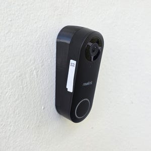 May include: Black Reolink video doorbell mounted on a white wall. The doorbell has a camera lens, a button, and the brand name "reolink" printed on it. A white sticker with text is affixed to the side.