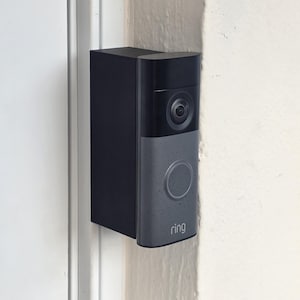 May include: A black and gray Ring video doorbell mounted on a white wall. The doorbell has a camera lens, a circular button, and the Ring logo. The device is rectangular with rounded edges.