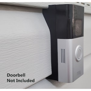 May include: A black and silver Ring doorbell mount attached to a white siding. The doorbell is not included.
