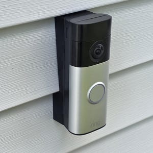 May include: A rectangular video doorbell with a black upper section containing a camera lens and a silver lower section featuring a circular button and the word ring printed at the bottom. The device is mounted vertically on light gray siding.