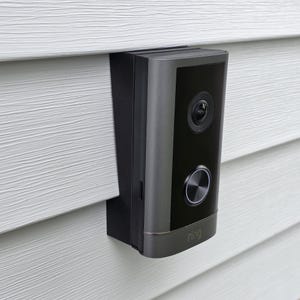 May include: A black and gray Ring video doorbell mounted on white siding. The doorbell has a camera lens, a button, and the Ring logo. The device is rectangular with rounded edges.