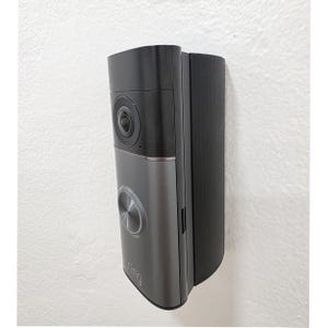 May include: A dark gray and black Ring video doorbell mounted on a white textured wall. The doorbell has a camera lens, a silver button, and the Ring logo. The device is rectangular with rounded edges.