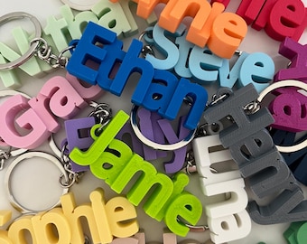 May include: A collection of colorful, personalized keychains with names like "Annie", "Ethan", "Jamie", "Chloe", and "Daniel". Each keychain features a 3D printed name in a different color, attached to a silver key ring.