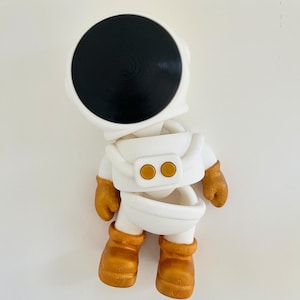 Puede incluir: Una figura de astronauta blanca con una visera de casco negra y guantes y botas doradas. El traje del astronauta tiene dos botones dorados. La figura es una representación caprichosa de la exploración espacial.