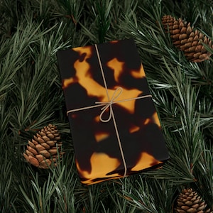 May include: A gift box wrapped in brown and orange tortoiseshell patterned paper with a brown twine bow. The box is surrounded by green pine branches and pine cones.