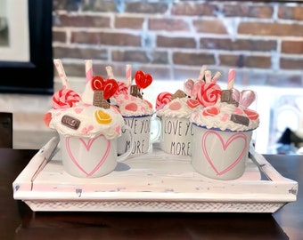May include: A white tray holds four decorative mugs with a pink heart design. Each mug is topped with faux whipped cream, candies, and a straw. The mugs have the words "LOVE YOU MORE" printed on them.
