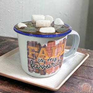 Puede incluir: Taza de cerámica blanca con borde azul y diseño de otoño. La taza está llena de un líquido marrón y cubierta con malvaviscos. La taza está sobre un platillo blanco. El texto en la taza dice "Fall is my favorite".