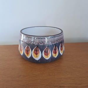 May include: A ceramic bowl with a dark gray exterior and a white interior. The bowl is decorated with a repeating pattern of stylized feather shapes in yellow, orange, and brown. The rim has a textured, light brown band.