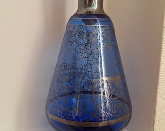May include: A cobalt blue glass decanter with a silver stopper and intricate silver detailing. The decanter has a wide base and a narrow neck.