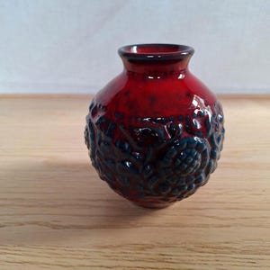 May include: A small, round ceramic vase with a glossy red glaze. The lower portion features a textured, dark blue floral design. The vase has a narrow neck and sits on a wooden surface. A decorative piece for home decor.