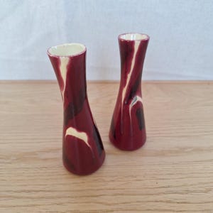 Könnte beinhalten: Zwei kleine, dekorative Vasen mit einem marmorierten Design in Burgunderrot und Creme. Die Vasen haben eine schlanke, geschwungene Form und einen cremefarbenen Innenraum. Sie stehen auf einer hellen Holzoberfläche.