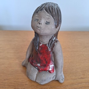 Pode incluir: Estatueta de cerâmica de uma menina sentada com cabelo comprido e escuro e olhos fechados. Ela usa um top vermelho e tem um corpo cinza. A estatueta está em uma superfície de madeira.