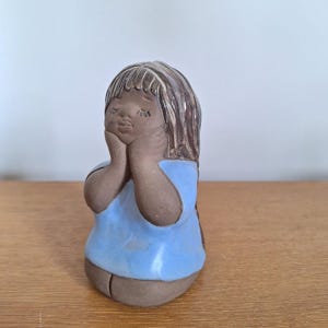 May include: A ceramic figurine of a girl with closed eyes, resting her chin on her hands. The figurine has brown hair and skin, and wears a light blue top. The piece is on a wooden surface.