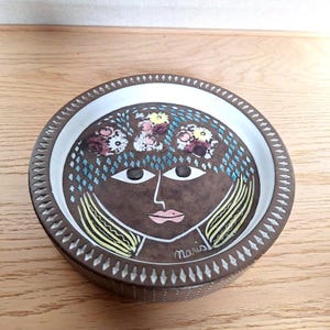 May include: A round, brown ceramic dish with a white rim and a decorative face design. The face has large eyes, a pink mouth, and yellow hair. The design includes floral accents and the name "Maris" is written on the side.