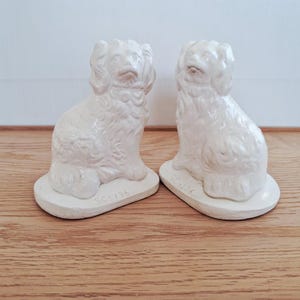 Op de afbeelding: Twee crèmekleurige keramische hondenbeeldjes. Elke hond zit op een kleine, ovale basis. De honden hebben gedetailleerde kenmerken, waaronder floppy oren en een halsband. De basis van elk beeldje heeft de tekst "SC-136".