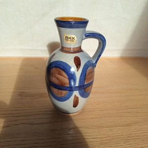 May include: A ceramic pitcher with a handle, featuring a blue rim and handle, with a light gray body. The pitcher is decorated with brown and blue circular and oval patterns. The label reads "BAY KERAMIK".