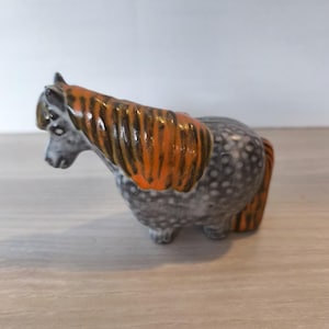 May include: A ceramic horse figurine featuring a dapple gray body with white spots and a vibrant orange mane and tail. The horse is depicted in a standing position with its head slightly lowered, showcasing a glossy finish and detailed texture.