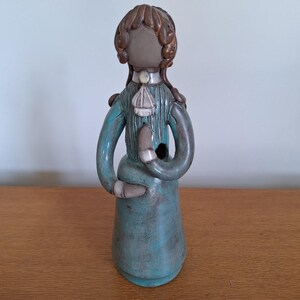 May include: A ceramic figurine of a person in a long teal dress with long sleeves. The figure has brown curly hair and a white collar. The hands are clasped in front, and the figure has a hole in the chest.