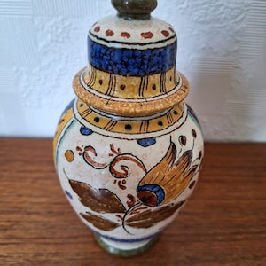 May include: A ceramic jar with a lid, decorated with a floral pattern in shades of brown, yellow, blue, and white. The jar has a green base and a green lid.