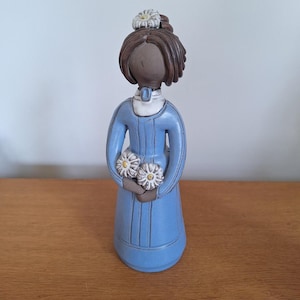 May include: A ceramic figurine of a woman in a long, pale blue dress, holding two white and yellow flower bouquets. The figurine has brown hair and a white flower on top of her head. The figure is standing on a wooden surface.