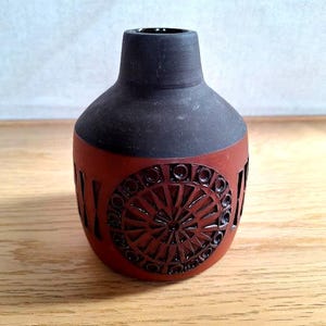 May include: A ceramic vase with a dark grey neck and a reddish-brown body. The body features a circular, geometric design with cut-out shapes. The vase has a rounded shape and a small opening at the top.