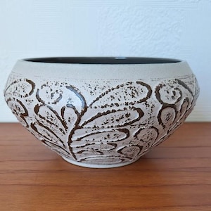 Puede incluir: Una maceta de cerámica blanca con un diseño floral marrón. La maceta tiene una forma redondeada y un borde ancho.