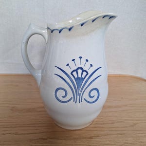 Puede incluir: Jarra de cerámica blanca con un diseño floral azul. La jarra tiene un asa curva y un pico. El diseño floral está centrado en la parte delantera e incluye una flor estilizada y detalles en espiral. El borde está decorado con guiones azules.