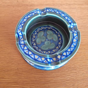 May include: A round ceramic ashtray with a blue and teal color scheme. The rim features a repeating pattern of blue triangles and yellow dots. The interior has a floral design in shades of blue and teal.
