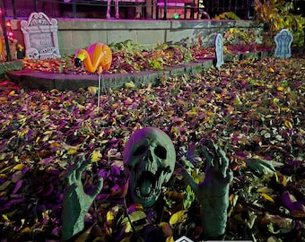 May include: A Halloween yard display featuring a zombie skull and hands emerging from a bed of autumn leaves. An orange flamingo decoration and decorative tombstones are also visible. The scene is lit with purple and orange lights.