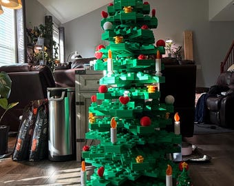 May include: A large green Christmas tree constructed from interlocking toy bricks, adorned with red ornaments, yellow star-shaped decorations, and white candle-shaped lights. A smaller, pre-made Christmas tree is also present.