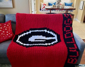 May include: A large, red knitted blanket with a black and white design. The blanket features a large "G" logo in the center and the word "DOGS" in black on the side. The blanket is draped over a gray sofa.
