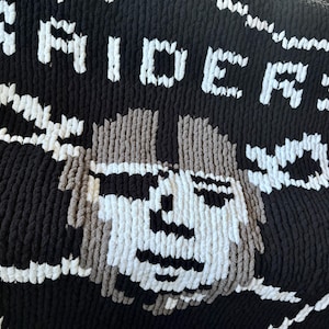 May include: Black and white knitted blanket with the word "RIDERS" in white yarn and a gray and white knitted image of a person's face.